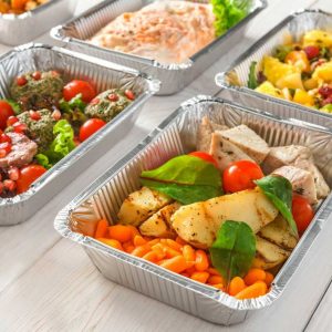 El menú diario, que incluye recipientes de aluminio para llevar con una variedad de comidas, está ordenado de forma ordenada sobre una superficie de madera blanca. La selección incluye platos como pollo a la parrilla con verduras, pescado sazonado y una mezcla de ensalada de frutas. Algunos recipientes están adornados con tomates cherry y verduras.