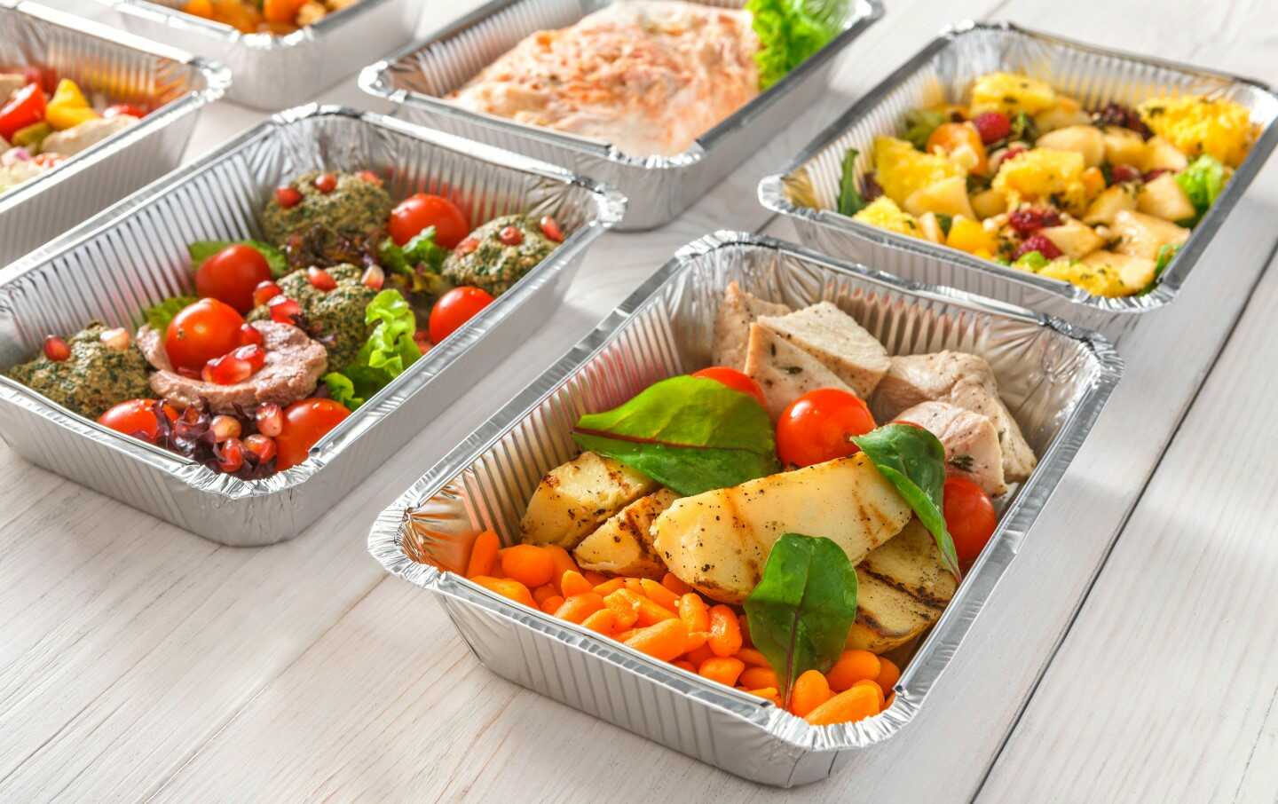 El menú diario, que incluye recipientes de aluminio para llevar con una variedad de comidas, está ordenado de forma ordenada sobre una superficie de madera blanca. La selección incluye platos como pollo a la parrilla con verduras, pescado sazonado y una mezcla de ensalada de frutas. Algunos recipientes están adornados con tomates cherry y verduras.